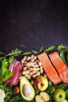 Fresh vegetables, fruits, fish, meat, nuts on black chalk board background. Сauliflower, avocado, apples, tomatoes, salmon, beef, spinach, herbs. Diet/healthy/paleo food. Ingredients. Space for text