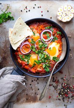 Shakshuka with pita bread in a pan. Middle eastern traditional dish. Fried eggs with tomatoes, bell pepper, vegetables and herbs. Shakshouka on a table. Top view. Sunny side up eggs. From above