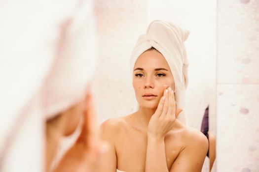 Young beautiful woman using face cream moisturizing lotion after bath. Pretty attractive girl in a towel on her head stands in front of a mirror in a home bathroom. Daily hygiene and skin care.