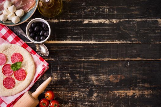 Pizza making background. Ingredients for making pizza. Space for text. Pizza dough, flour, cheese, mozzarella, tomatoes, basil, pepperoni, olives and rolling pin over black wooden background. Top view