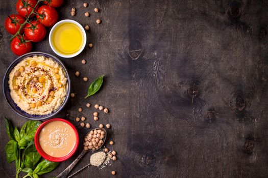 Bowl with hummus, chickpea, tahini, olive oil, sesame seeds, cherry tomatoes and herbs on dark rustic wooden background. Space for text. Food frame. Middle eastern cuisine. Top view. Hummus background