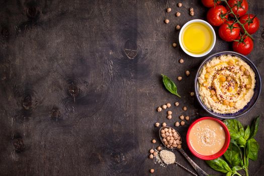 Bowl with hummus, chickpea, tahini, olive oil, sesame seeds, cherry tomatoes and herbs on dark rustic wooden background. Space for text. Food frame. Middle eastern cuisine. Top view. Hummus background