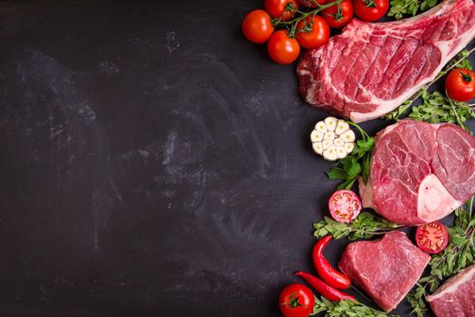 Raw juicy meat steaks ready for roasting on a black chalk board background. Rib eye steak on the bone, veal shank (ossobuco), fillet with cherry tomatoes, hot pepper and herbs. Space for text