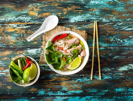 Traditional Vietnamese soup Pho bo with herbs, meat, rice noodles, broth. Pho bo in bowl with chopsticks, spoon. Top view. Asian soup Pho bo, wooden table background. Vietnamese authentic tasty soup