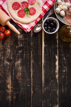 Pizza making background. Ingredients for making pizza. Space for text. Pizza dough, flour, cheese, mozzarella, tomatoes, basil, pepperoni, olives and rolling pin over black wooden background. Top view