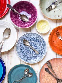 Different vintage multicolored empty plates and bowls on a white old wooden table. With antique spoons and forks. Table setting. Shabby chic/retro style. Top view. Rustic kitchen. Set of plates