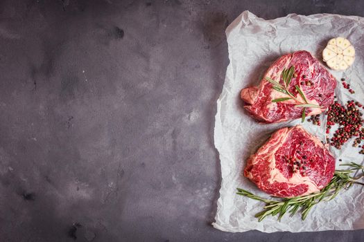 Raw juicy ribeye steaks with seasonings ready for roasting on baking paper. Rustic concrete background. Fresh marbled meat steaks with herbs, garlic, olive oil, pepper, salt. Space for text. Top view