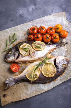 Tasty baked whole fish on baking paper. Baked sea bream with lemon, onion, herbs, cherry tomatoes, spices on dark rustic background. Grilled delicious fish. Diet and healthy food. Top view