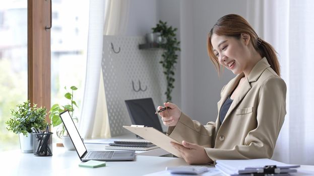 Attractive young woman sitting and her workplace and checking information on clipboard.