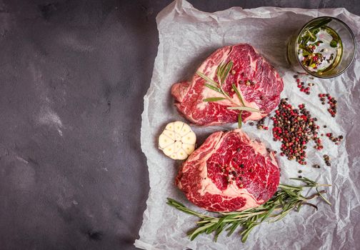 Raw juicy ribeye steaks with seasonings ready for roasting on baking paper. Rustic concrete background. Fresh marbled meat steaks with herbs, garlic, olive oil, pepper, salt. Space for text. Top view