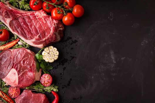 Raw juicy meat steaks ready for roasting on a black chalk board background. Rib eye steak on the bone, veal shank (ossobuco), fillet with cherry tomatoes, hot pepper and herbs. Space for text