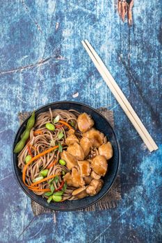 Asian noodles with chicken, vegetables, bowl, rustic wooden blue background. Closeup. Top view. Soba noodles, teriyaki chicken, edamame, chopsticks. Asian style dinner/lunch. Chinese/Japanese noodles