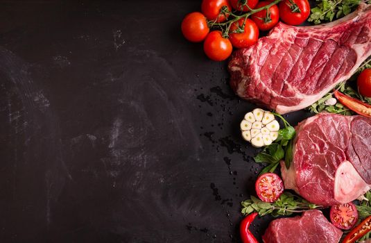 Raw juicy meat steaks ready for roasting on a black chalk board background. Rib eye steak on the bone, veal shank (ossobuco), fillet with cherry tomatoes, hot pepper and herbs. Space for text