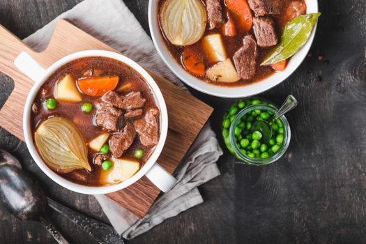 Meat stew with beef, potato, carrot, pepper, spices, green peas. Slow cooked meat stew, bowl, wooden background. Hot autumn/winter dish. Closeup. Top view. Comfort food. Homemade soup/ragout/casserole