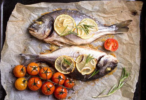 Tasty baked whole fish on baking paper. Baked sea bream with lemon, onion, herbs, cherry tomatoes, spices on dark rustic background. Grilled delicious fish. Diet and healthy food. Top view