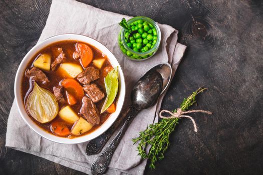 Meat stew with beef, potato, carrot, onion, spices. green peas. Slow cooked meat stew in bowl, wooden background. Hot autumn/winter dish. Closeup. Top view. Space for text. Comfort food. Homemade soup