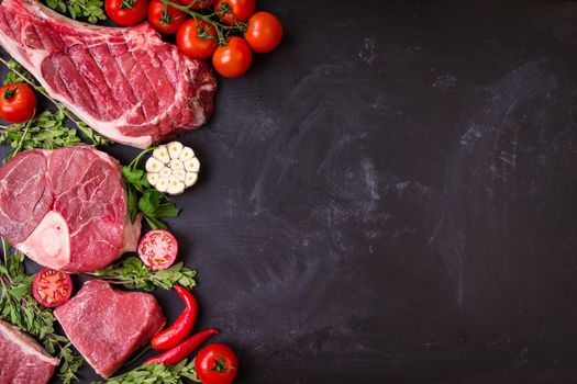 Raw juicy meat steaks ready for roasting on a black chalk board background. Rib eye steak on the bone, veal shank (ossobuco), fillet with cherry tomatoes, hot pepper and herbs. Space for text