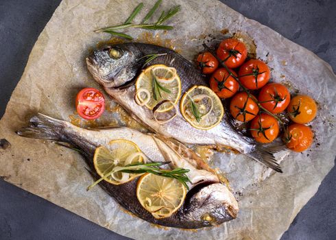 Tasty baked whole fish on baking paper. Baked sea bream with lemon, onion, herbs, cherry tomatoes, spices on dark rustic background. Grilled delicious fish. Diet and healthy food. Top view