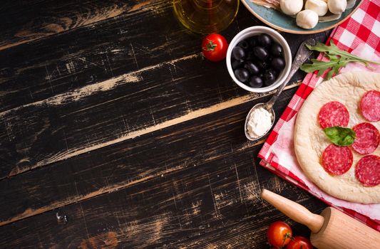 Pizza making background. Ingredients for making pizza. Space for text. Pizza dough, flour, cheese, mozzarella, tomatoes, basil, pepperoni, olives and rolling pin over black wooden background. Top view