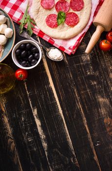 Pizza making background. Ingredients for making pizza. Space for text. Pizza dough, flour, cheese, mozzarella, tomatoes, basil, pepperoni, olives and rolling pin over black wooden background. Top view