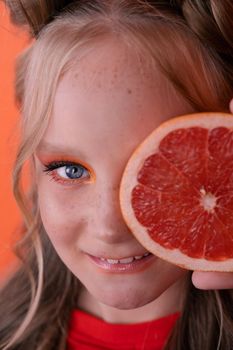 young girl with grapefruit slice on orange background. curly hair and two ponytails.