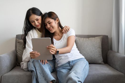 Happy young lesbian couple hugging having fun using digital tablet relaxing on couch at home. Two smiling asian women friends holding computer looking at screen enjoying surfing online watching videos.
