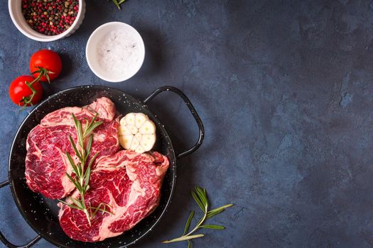 Raw juicy steaks with seasonings in a pan ready for roasting on rustic concrete background. Fresh marbled meat steaks with herbs, garlic, olive oil, pepper, salt and tomatoes. Space for text. Top view