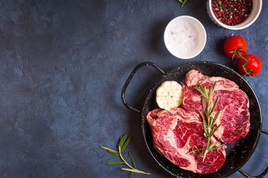 Raw juicy steaks with seasonings in a pan ready for roasting on rustic concrete background. Fresh marbled meat steaks with herbs, garlic, olive oil, pepper, salt and tomatoes. Space for text. Top view