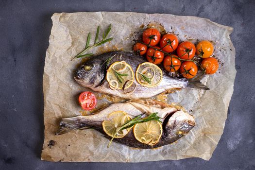 Tasty baked whole fish on baking paper. Baked sea bream with lemon, onion, herbs, cherry tomatoes, spices on dark rustic background. Grilled delicious fish. Diet and healthy food. Top view