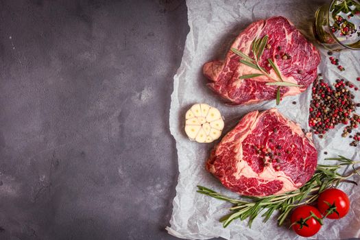 Raw juicy ribeye steaks with seasonings ready for roasting on baking paper. Rustic concrete background. Fresh marbled meat steaks with herbs, garlic, oil, spices, tomatoes. Space for text. Top view