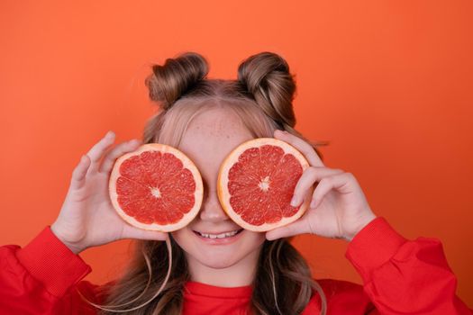 young girl with grapefruit slice on orange background. curly hair and two ponytails.