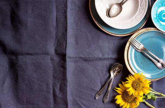 Vintage multicolored empty plates and bowls on a dark gray linen tablecloth with sunflower. With antique spoons and forks. Space for text. Table setting. Shabby chic/retro style. Top view. Copy space