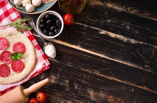 Pizza making background. Ingredients for making pizza. Space for text. Pizza dough, flour, cheese, mozzarella, tomatoes, basil, pepperoni, olives and rolling pin over black wooden background. Top view