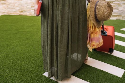 cropped shot of a young woman, entering the hotel reception with her suitcase on wheels. woman on a pleasure trip dressed in summer clothes. Outside, dim sunlight, grass floor with white tiles.