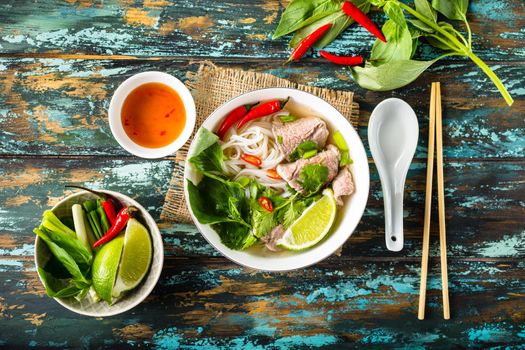 Traditional Vietnamese soup Pho bo with herbs, meat, rice noodles, broth. Pho bo in bowl with chopsticks, spoon. Space for text. Top view. Asian soup Pho bo on wooden table background. Vietnamese soup