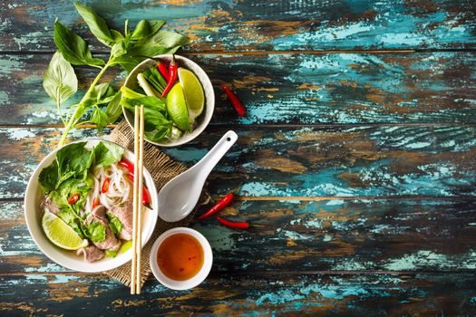 Traditional Vietnamese soup Pho bo with herbs, meat, rice noodles, broth. Pho bo in bowl with chopsticks, spoon. Space for text. Top view. Asian soup Pho bo on wooden table background. Vietnamese soup