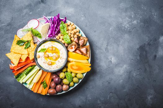 Hummus platter with assorted snacks, stone background. Space for text. Hummus, vegetables sticks, chickpeas, olives, pita chips. Plate, Middle Eastern/Mediterranean meze. Party/finger food. Top view