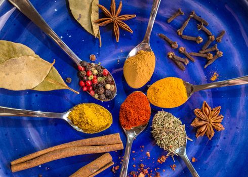 Different colorful spices in little spoons on dark blue background. Curry, turmeric, cinnamon sticks, bay leaves, sumac, cloves, peppercorn, paprika, rosemary, star anise. Top view. Closeup