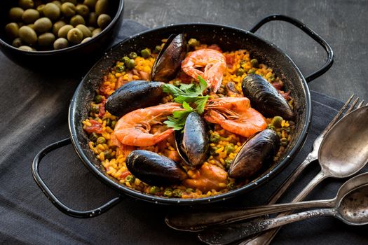 Paella in black pan with rice, shrimps, mussels, squid and meat, bowl with olives and vintage cutlery. Seafood paella, traditional spanish dish. Paella on rustic black wooden table. Selective focus
