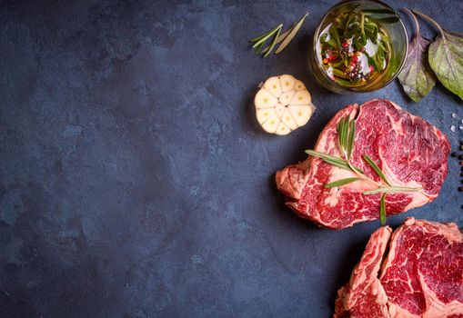 Raw juicy ribeye steaks with seasonings ready for roasting on rustic concrete background. Fresh marbled meat steaks with herbs, garlic, olive oil, whole pepper and rock salt. Space for text. Top view