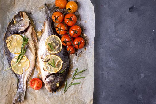 Tasty baked whole fish on baking paper. Baked sea bream with lemon, onion, herbs, cherry tomatoes, spices on dark rustic background. Grilled delicious fish. Diet and healthy food. Top view