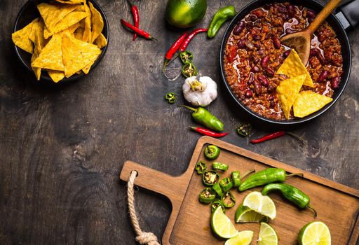 Chili con carne in frying pan on dark wooden background. Ingredients for making Chili con carne. Space for text. Top view. Chili with meat, nachos, lime, hot pepper. Mexican/Texas traditional dish