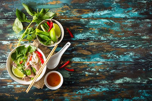 Traditional Vietnamese soup Pho bo with herbs, meat, rice noodles, broth. Pho bo in bowl with chopsticks, spoon. Space for text. Top view. Asian soup Pho bo on wooden table background. Vietnamese soup