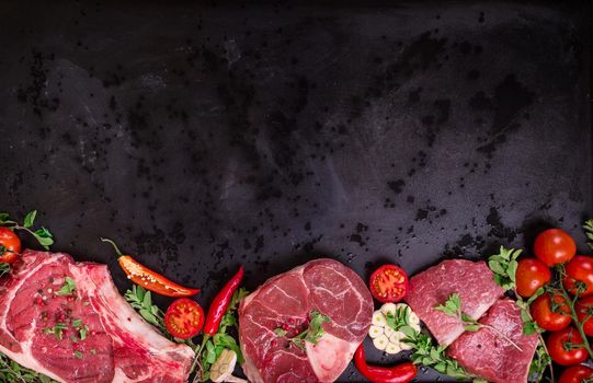 Raw juicy meat steaks ready for roasting on a black chalk board background. Rib eye steak on the bone, veal shank (ossobuco), fillet with cherry tomatoes, hot pepper and herbs. Space for text