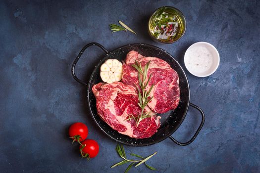 Raw juicy steaks with seasonings in a black pan ready for roasting on rustic concrete background. Fresh marbled meat steaks with herbs, garlic, olive oil, pepper, salt and tomatoes. Top view