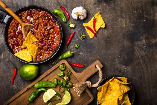 Chili con carne in frying pan on dark wooden background. Ingredients for making Chili con carne. Space for text. Top view. Chili with meat, nachos, lime, hot pepper. Mexican/Texas traditional dish