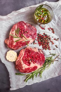 Raw juicy steaks with seasonings on a baking paper ready for roasting on rustic concrete background. Fresh marbled meat steaks with herbs, garlic, olive oil, colorful whole pepper, rock salt. Top view