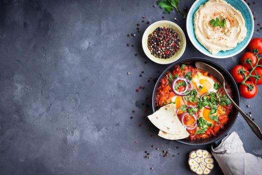 Shakshuka with pita bread in pan, hummus in bowl on rustic background. Middle eastern traditional dishes. Fried eggs with vegetables. Top view. Space for text. Middle eastern style breakfast or lunch