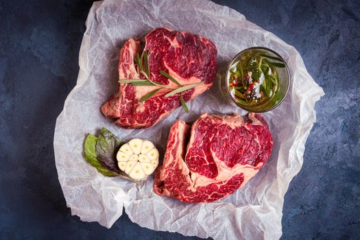 Raw juicy steaks with seasonings on a baking paper ready for roasting on rustic concrete background. Fresh marbled meat steaks with herbs, garlic, olive oil, colorful whole pepper, rock salt. Top view