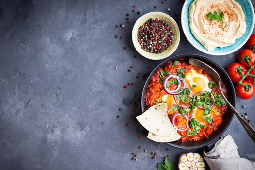 Shakshuka with pita bread in pan, hummus in bowl on rustic background. Middle eastern traditional dishes. Fried eggs with vegetables. Top view. Space for text. Middle eastern style breakfast or lunch